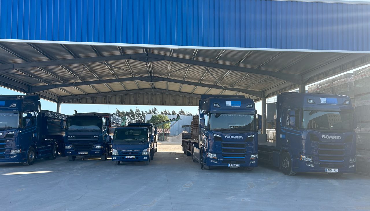 Cinco camiões da frota estacionados de frente, sob a cobertura metálica azul do parque