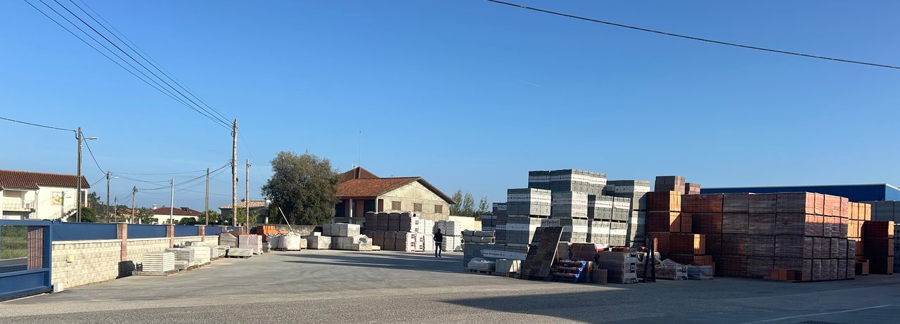 Vista alargada do parque, com paletes de tijolo e bloco empilhadas ao fundo, casas e linhas elétricas atrás